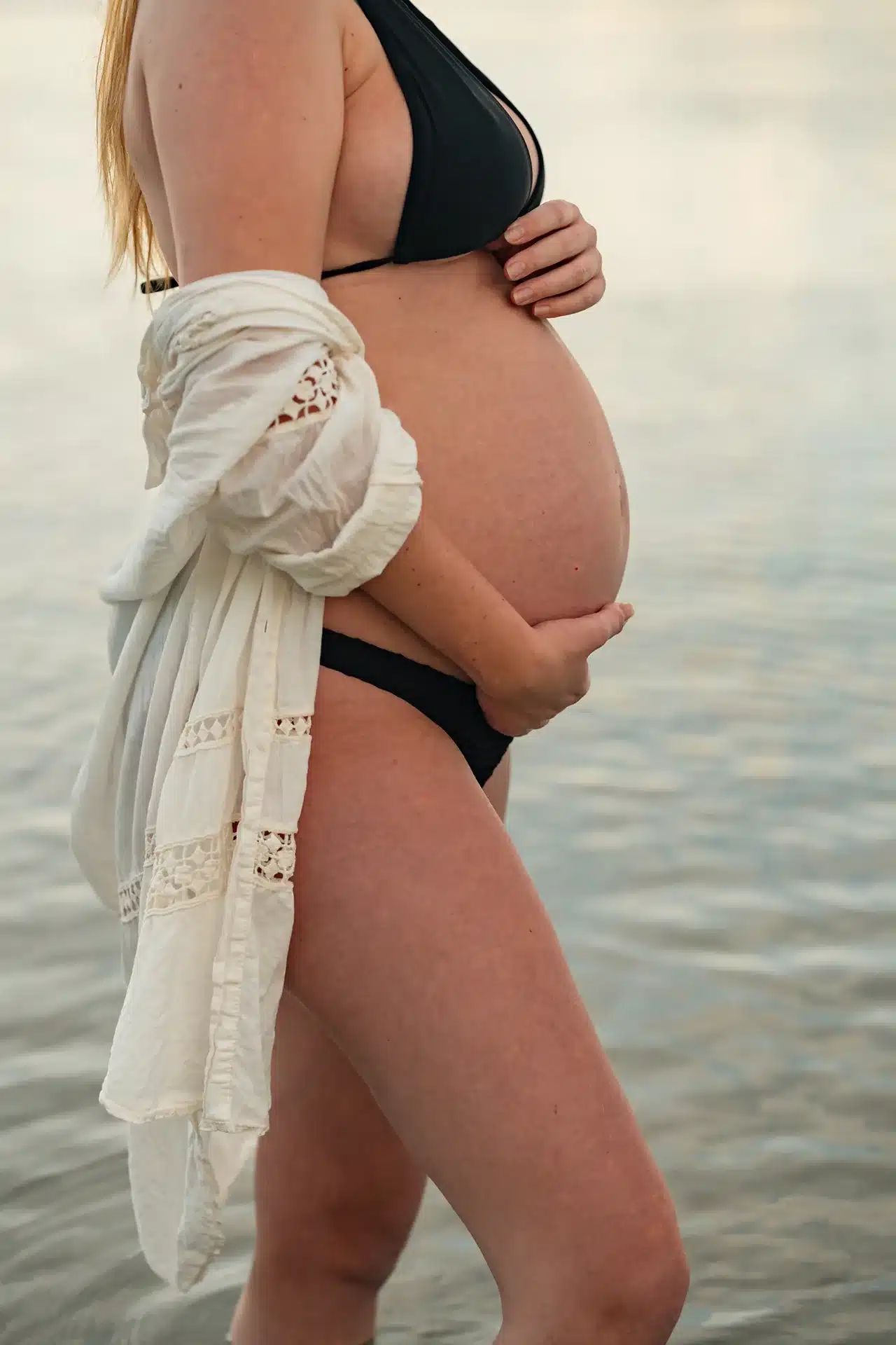 Shooting grossesse à Tahiti avec une future maman qui tient son ventre les pieds dans l'eau